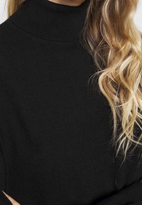 Pull noir à col roulé avec une texture douce, doté d'un col haut et de manches longues, partiellement caché par des cheveux blonds dorés, lâches et ondulés.