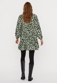 Robe fleurie verte avec motifs blancs, design à volants, manches longues et forme évasée ; assortie à des leggings noirs et des bottes montantes.