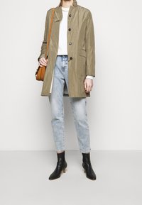Personne portant un trench-coat marron clair, un haut blanc, un jean délavé, des bottines noires et un sac à épaule marron sur un fond uni.