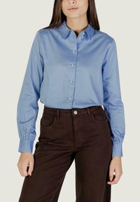 Donna che indossa una camicia azzurra chiara con bottoni e maniche lunghe, insieme a pantaloni marroni a vita alta, che sostiene contro uno sfondo neutro.