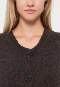 Brun, strukturert cardigan med rund hals og to svarte, runde knapper foran. Fremhever varme og avslappet stil.