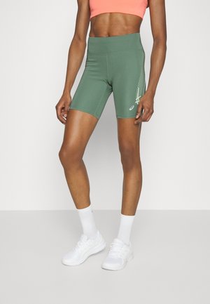 Athletische Person trägt grüne hoch taillierte Fahrradshorts mit seitlichem Design, korallenfarbenes kurzes Oberteil, weiße Socken und weiße Laufschuhe.