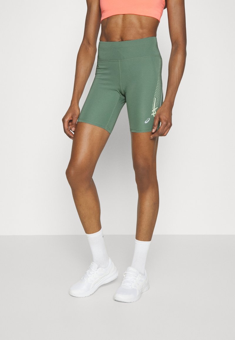 Personne athlétique portant un short de vélo vert taille haute avec un motif sur le côté, un crop top corail, des chaussettes blanches et des chaussures de course blanches.