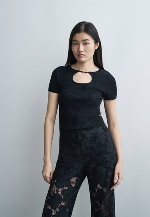 Noor naine seisab tavalise tausta ees, kandes musta lühikeste varrukatega topi avaga kaelas ja musti pitspükse.