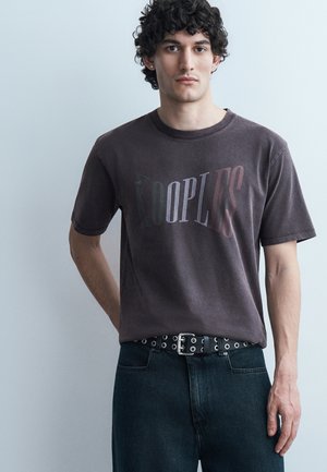 Jonge man met krullend haar, gekleed in een zwart verweerd The Kooples T-shirt, ingestopt in een donkere spijkerbroek met zwarte riem, staand tegen een effen lichte muur.