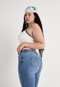 White tank top with black trim paired with light blue denim jeans. Model has long curly hair and is wearing a white bandana.
