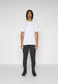 Witte sportt-shirt met groene en gele accenten, gecombineerd met donkergrijze broek met gele strepen aan de zijkanten. Zwarte schoenen.