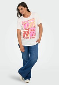 Hvid t-shirt med farverigt Rolling Stones lip print, der har fire variationer af pink og orange læber. Sæt sammen med blå flared jeans.