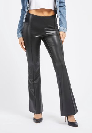 Frau trägt schwarze hoch taillierte Kunstleder-Flared-Hose, schwarze spitze High Heels, weißes kurzes Top und Jeansjacke und steht vor weißem Hintergrund.