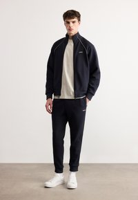 Νέος άνδρας με μαύρη φόρμα BOSS και λευκά αθλητικά παπούτσια, στέκεται μπροστά σε ένα απλό ανοιχτόχρωμο τοίχο και δάπεδο από σκυρόδεμα.