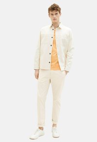 Lys beige knapfyldt jakke over en orange skjorte, parret med cremefarvede bukser og hvide sneakers, med to brystlommer.