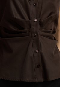Brun knapop skjorte med en samlet front, der har blanke knapper og en glat tekstur, designet med subtile plisseringer for ekstra detaljer.