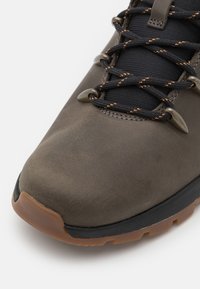 Zapato de senderismo de cuero marrón con puntera redondeada, lengüeta de malla negra y cordones con acentos naranjas. Parte superior texturizada y suela de goma para agarre.