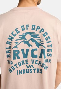 Lyse pink bomulds T-shirt med blå typografisk tryk med teksten "Balance of Opposites RVCA Nature Versus Industry." Korte ærmer.