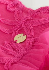 Rosa Netzstoff mit Spitzenband-Details und einem goldfarbenen Metalleinsatz mit einem geprägten "B"-Logo. Strukturierte Schichten erzeugen visuelles Interesse.