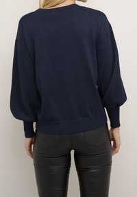 Marineblauer Strickpullover mit strukturierter Oberfläche, ausgestattet mit Ballonärmeln und einem gerippten Saum. Kombiniert mit schwarzen, figurbetonten Hosen.