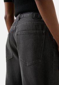 Czarne spodnie dżinsowe o luźnym kroju, z dwoma tylnymi kieszeniami i czarnym okrągłym guzikiem z logo "B" na pasku.