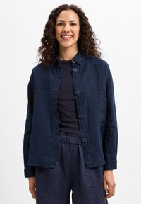 Navy blue linen shirt with button closure, a patch pocket, and a relaxed fit. Worn over a dark top, paired with matching trousers.