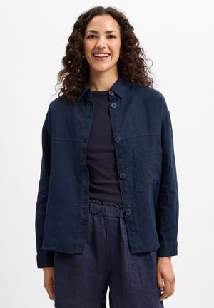 Navy blue linen shirt with button closure, a patch pocket, and a relaxed fit. Worn over a dark top, paired with matching trousers.