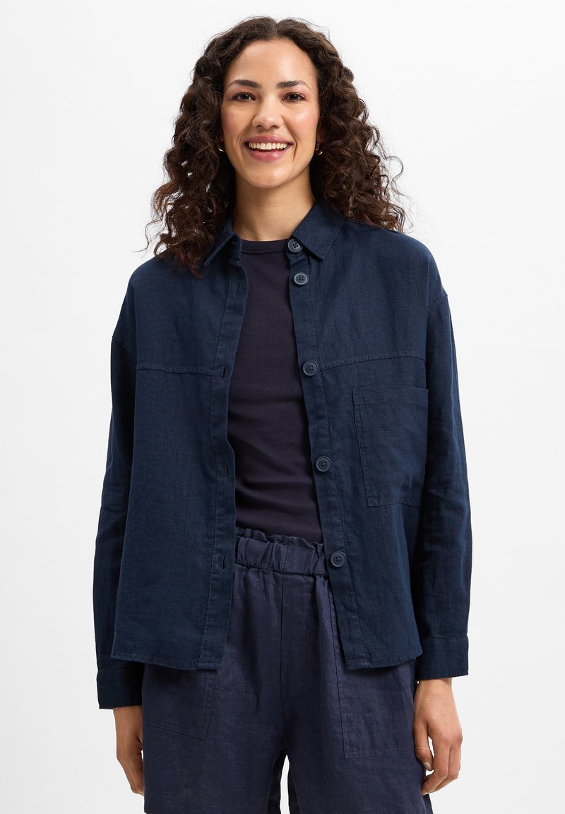 Navy blue linen shirt with button closure, a patch pocket, and a relaxed fit. Worn over a dark top, paired with matching trousers.