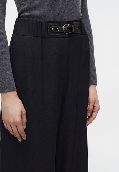 Gros plan d'une personne portant un pantalon noir taille haute avec une ceinture noire et un haut gris côtelé à manches longues, les mains reposant le long du corps.