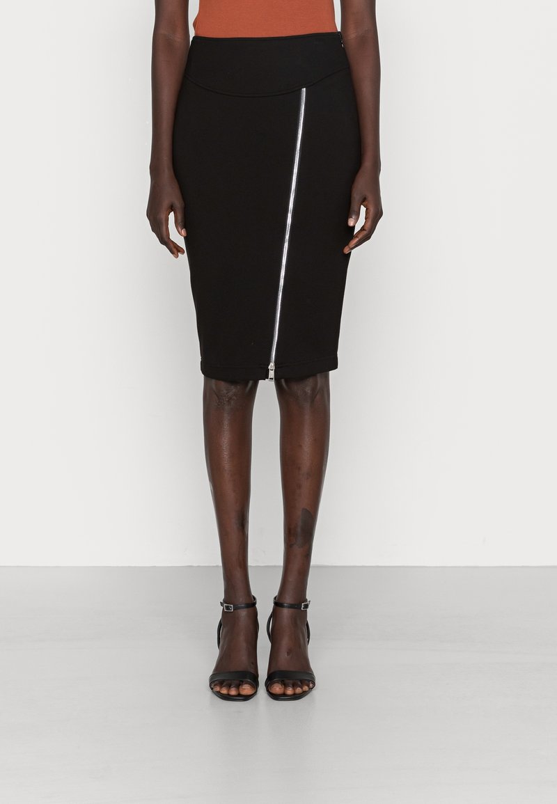 Jupe crayon noire mi-longue avec fermeture éclair asymétrique argentée à l'avant, portée avec des sandales à talons hauts noires à bride cheville.