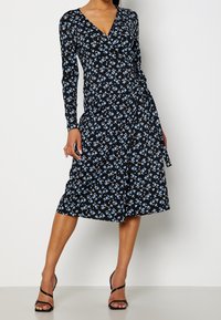 Musta pitkähihainen mekko, jossa on kietaisumalli ja sininen kukkakuosi. Sileä kangas, polveen ulottuva hame ja sidottu vyötäröyksityiskohta.