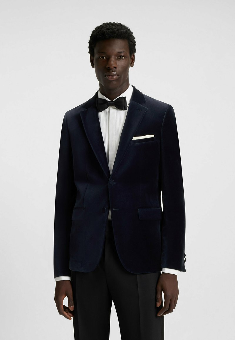 Giacca da smoking in velluto blu scuro con due tasche frontali e revers in satin, indossata sopra una camicia bianca plissettata e un papillon nero.