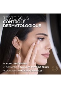 Application d'un produit de soin de la peau avec une personne touchant sa joue. Texture lisse, légère et lumineuse. Teint neutre, boucles d'oreilles en or simple.