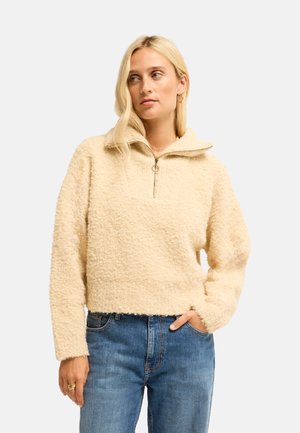 Donna che indossa un pullover in pile beige texturizzato con collo a mezzo zip e jeans blu, in piedi con una mano in tasca.