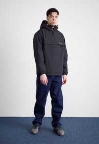 Black lightweight pullover jacket with a front pocket, elastic cuffs, and a hood. Paired with dark blue jeans and gray sneakers.