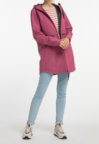 Chaqueta impermeable rosa con capucha, cremallera frontal y dos bolsillos laterales. Combinada con vaqueros azul claro y zapatillas multicolores.
