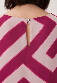 Elegante, leichte Bluse mit geometrischem Muster in Dunkelrosa und Hellbeige, mit Schlüsselöffnung und Metalknopf-Detail auf der Rückseite.