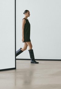 Czarna sukienka bez rękawów, czarne wysokie skórzane kozaki do kolan na płaskiej podeszwie. Minimalistyczny design, gładka faktura, stojąca na polerowanej betonowej podłodze.