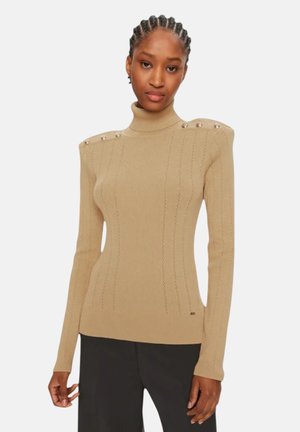 Donna con capelli intrecciati indossa un maglione a collo alto beige a coste con bottoni sulle spalle e pantaloni neri su uno sfondo bianco uniforme.