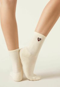 Legs wearing cream ribbed socks with small heart-shaped patch featuring text and decorative elements on outer ankle against a neutral background.