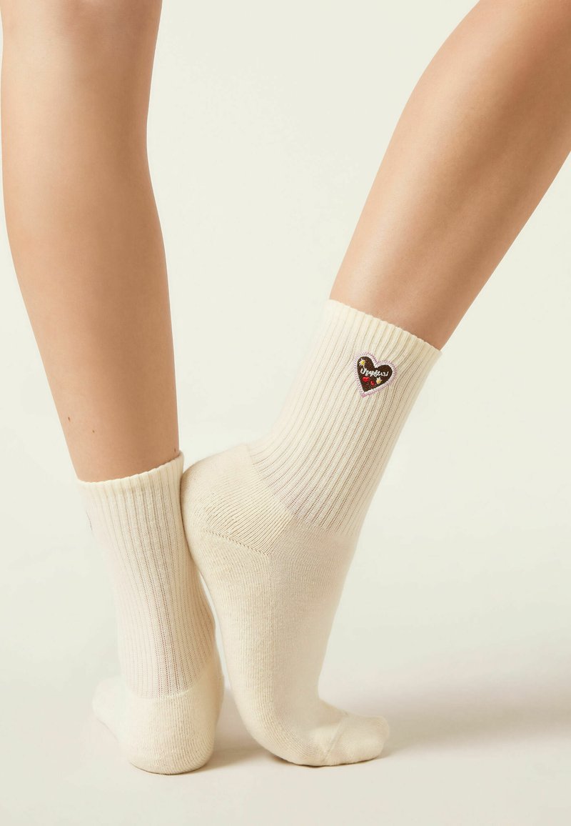 Legs wearing cream ribbed socks with small heart-shaped patch featuring text and decorative elements on outer ankle against a neutral background.