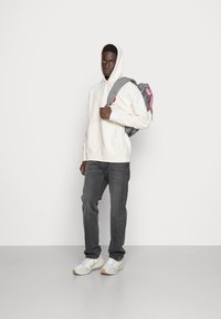 Zaino grigio con dettagli rosa indossato sopra una felpa bianca e jeans scuri. Le sneakers sono bianche con dettagli grigi chiari e beige.