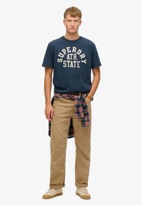 Camiseta de algodón azul oscuro con "SUPERDRY ATH. STATE" impresa en blanco. Pantalones de pana beige y una camisa de franela a cuadros atada en la cintura. Zapatillas tan.