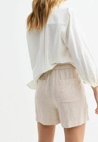 Žena v volné bílé košili s dlouhým rukávem a béžových lněných šortkách s kapsami na zadní straně, stojící na jednobarevném bílém pozadí.