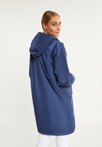 Jachetă impermeabilă de culoare albastru marin, cu glugă, mâneci lungi și o croială lejeră. Dispune de buzunare laterale și o textură netedă, strălucitoare.