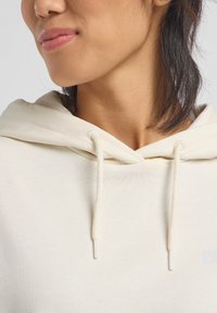 Felpa leggera color beige con cappuccio e stringhe, in tessuto morbido, con una texture a coste e un design casual. Presenta un piccolo logo nell'angolo.