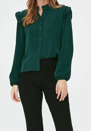 Vrouw draagt een donkergroene blouse met knopen, gerimpelde schouders en lange mouwen, gecombineerd met zwarte nauwsluitende broek.