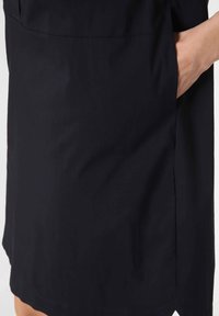 En persons hand vilar i sidofickan på ett knä-långt svart plagg mot en vit bakgrund.