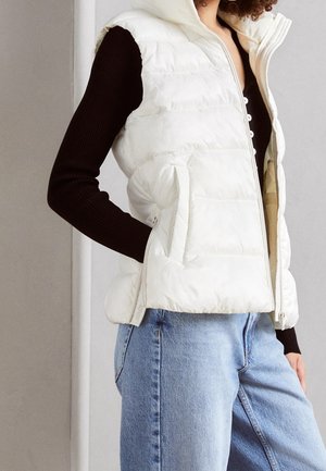 Persoon draagt een wit gewatteerd mouwloos vest over een zwart shirt met lange mouwen en lichtblauwe spijkerbroek, met hand in het vestzakje, staand binnen.