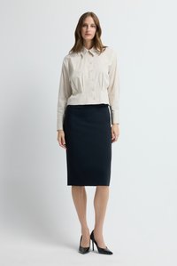 Kvinna stående iförd en beige skjorta med knappar, svart pennkjol till knäna och svarta högklackade skor mot en enfärgad bakgrund.