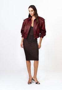 Kobieta ubrana w bordową skórzaną kurtkę bombową na dopasowanej czarnej przezroczystej sukience midi i czarnych sandałach na wysokim obcasie, stojąca na tle białym.