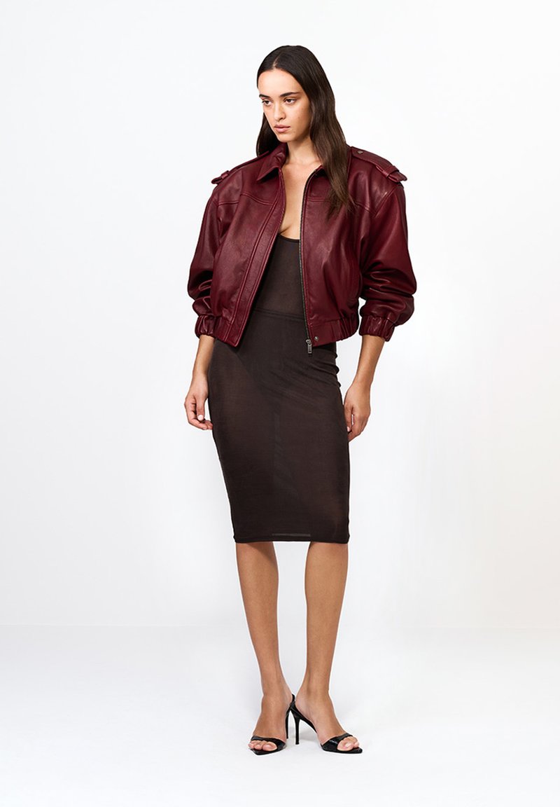 Kobieta ubrana w bordową skórzaną kurtkę bombową na dopasowanej czarnej przezroczystej sukience midi i czarnych sandałach na wysokim obcasie, stojąca na tle białym.