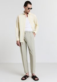 Uomo in camicia giallo chiaro sopra una canottiera bianca, pantaloni grigi chiari, sandali neri e occhiali da sole, in piedi davanti a uno sfondo chiaro uniforme.