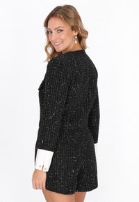 Chaqueta de tweed negra con acentos metálicos, detalles en los puños blancos y un corte entallado, llevada por un modelo, mostrando la vista trasera.